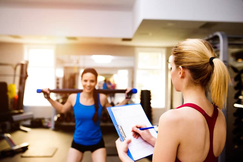 Woman in blue singlet exercising squats with bar in a gym with her personal trainer writing into exercise plan on clipboard