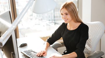 Photo of joyful young lady worker using computer in office and typing by keyboard. Look at computer.