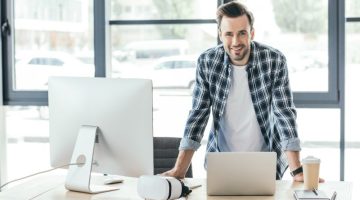 young man smiling at camera while leaning at table with laptop and desktop computer