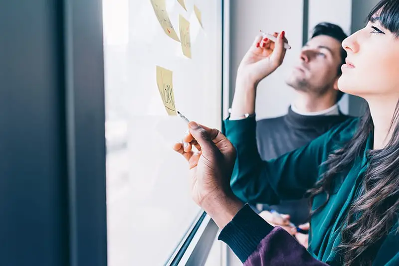 Three business people having a meeting in office. They are standing in front of glass wall with post it notes, pointing and discussing - business, teamwork, brainstorming concept
