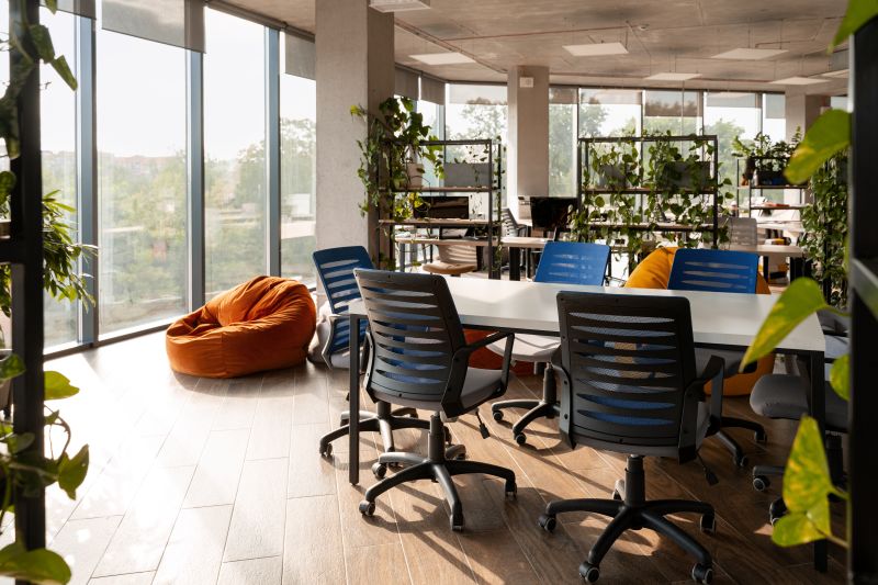 Sunlight streams into a modern coworking space, illuminating comfortable office chairs, beanbag chairs, and lush green plants, creating a welcoming and productive environment