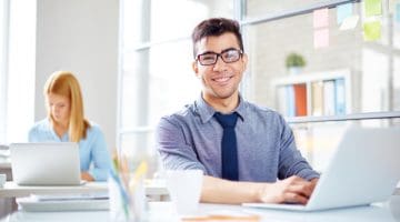 Young man sat at a desk using a laptop, female in the background using laptop