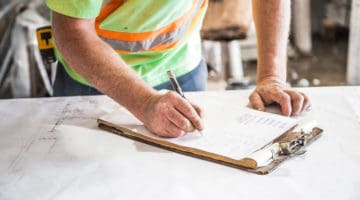Workman writing on a clipboard placed on top of blueprints