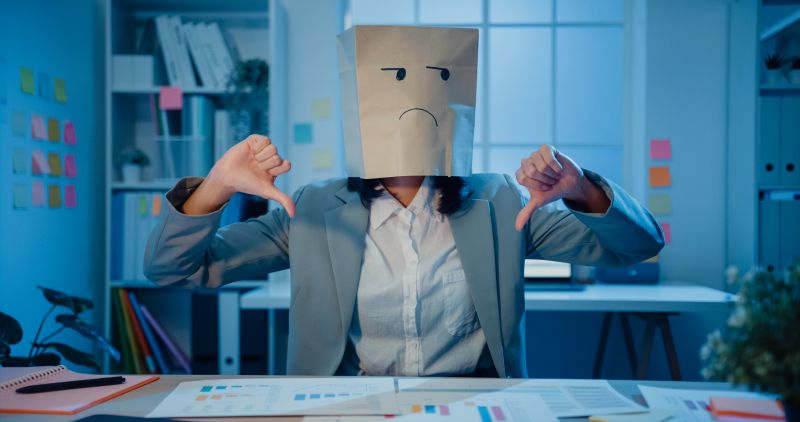 Young Asian businesswoman draw sad mask paper craft bag feel bad mood with fail work project.