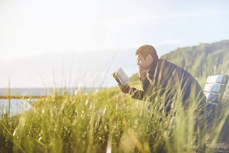 Man reading book on bench long grass sea view in background
