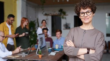 Woman standing in office wearing eyeglasses – with business colleagues behind her