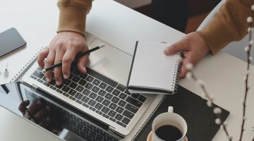 Person holding black pencil and notebook typing on laptop