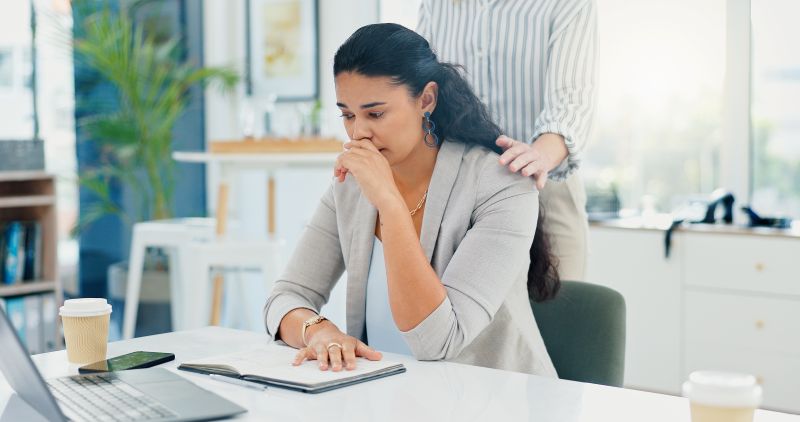 Worried woman sitting at an office desk with her hand near her mouth while a person standing behind her rests a hand on her shoulder.
