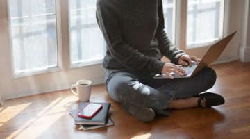 woman in grey sweater sitting cross legged on floor with laptop on knees