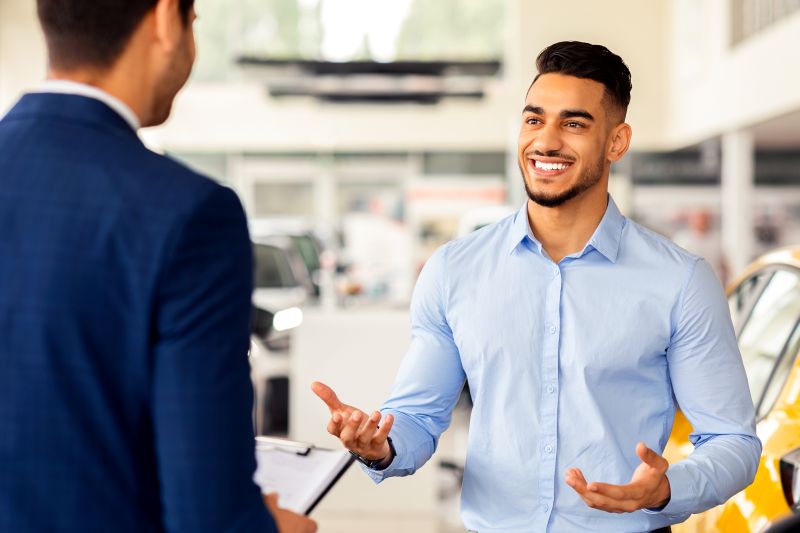 Smiling man in a light blue shirt talking and gesturing to a suited man holding a clipboard in a car showroom.