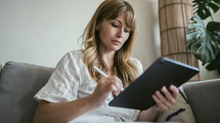 Woman using a stylus writing on a digital tablet while sitting in the couch