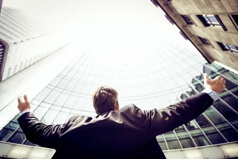 Man in suit outside looking up to building with arms raised signifying success