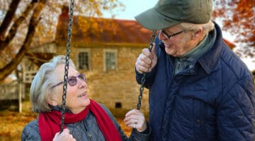 An old man standing beside old woman on swing