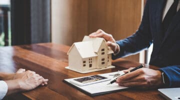 Two people in front of wooden table beside cartoon miniature house