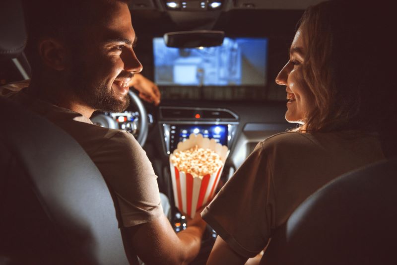 Beautiful happy young couple sitting in the car together and eating popcorn at a drive-in cinema