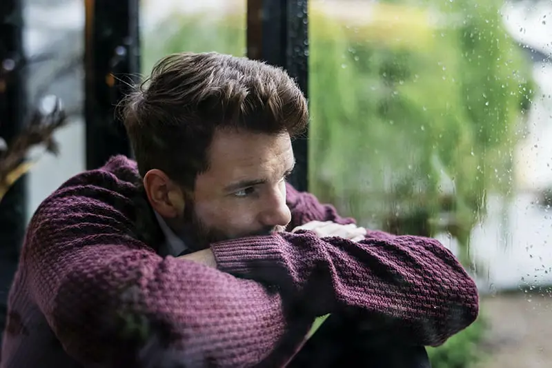 A sad man wearing maroon long sleeves near the window glass