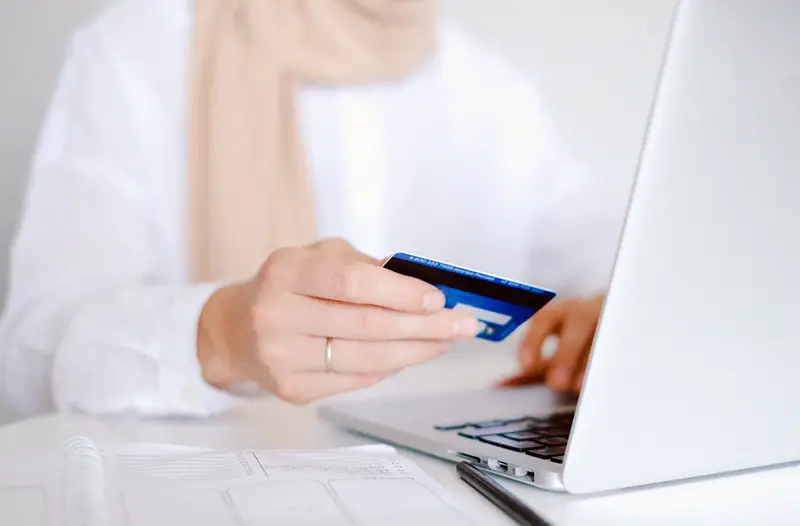 Person wearing white long sleeved shirt holding a credit card and making online payment on laptop