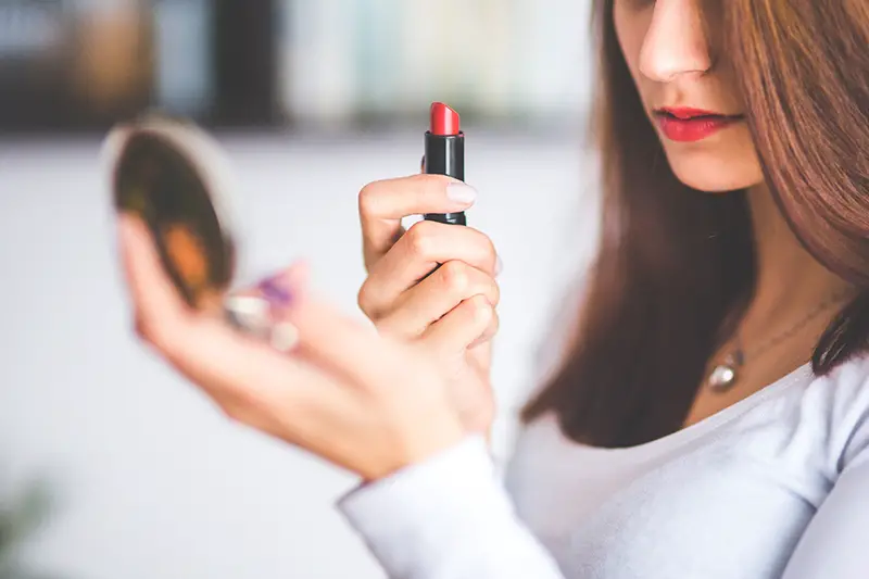 Woman with brown hair holding mirror while putting lipstick on