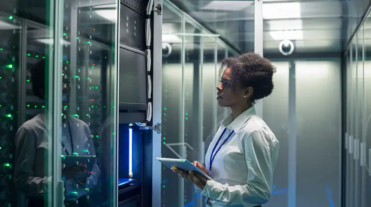 Female technician working on a tablet in a data center full of rack servers running diagnostics and maintenance on the system