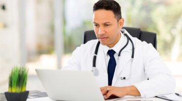 Doctor or medical consultant in white coat with and stethoscope sitting at a desk working on laptop