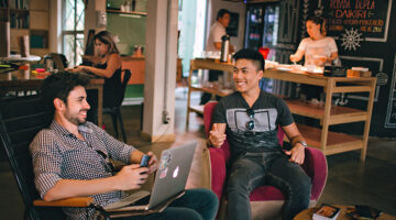 Men sitting in a coffee shop having a conversation and one working on a laptop