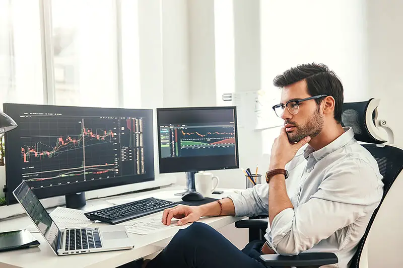 Young bearded trader wearing eyeglasses using his laptop while sitting in office in front of computer