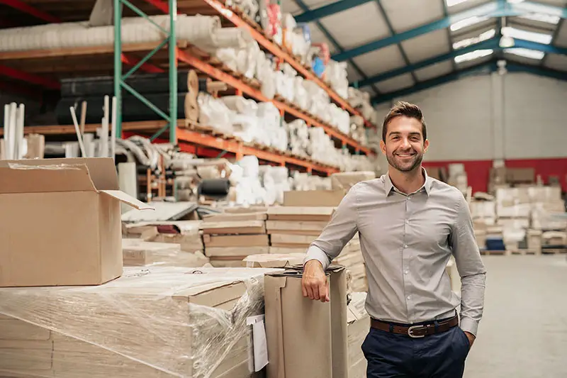 Portrait of a smiling warehouse manager leaning against some stock with piles of carpets stacked on shelves in the background