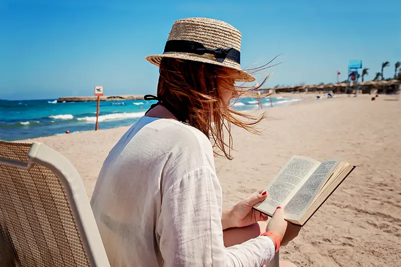 Young adult woman with a hat on the beach reading a book