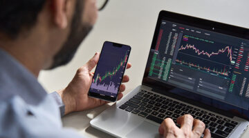 Business man trader investor analyst using mobile phone app analytics for cryptocurrency financial market analysis, trading data index chart graph on smartphone and laptop screen. Over shoulder view