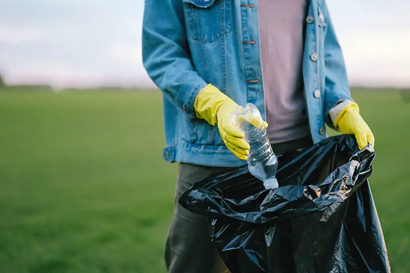 person in blue denim jacket putting plastic bottle inside a black garbage bag