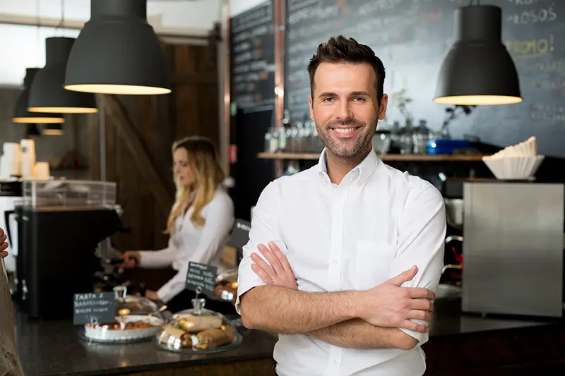 Male small business owner standing with crossed arms with employee in background preparing coffee