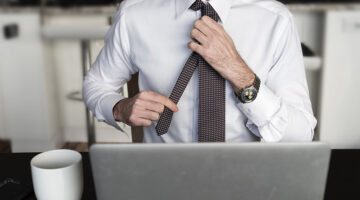 Headless man at desk adjusting his tie in front of laptop with coffee while working from home