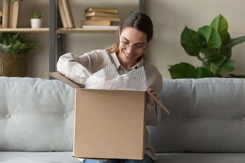 Smiling satisfied young woman customer sit on sofa unpack package open parcel