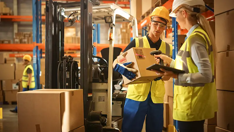 In Warehouse Manager Uses Digital Tablet and Scans Cardboard Boxes for Inventory, Talks with Forklift Driver about Package Delivery. Workers in Global Distribution Center with Shelves with Goods