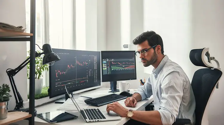 Young bearded trader in eyeglasses working with laptop while sitting in his modern office in front of computer screens with trading charts.