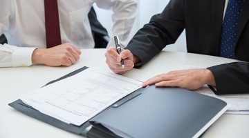 A Person in Black Suit Holding a Pen Near the Documents on the Table