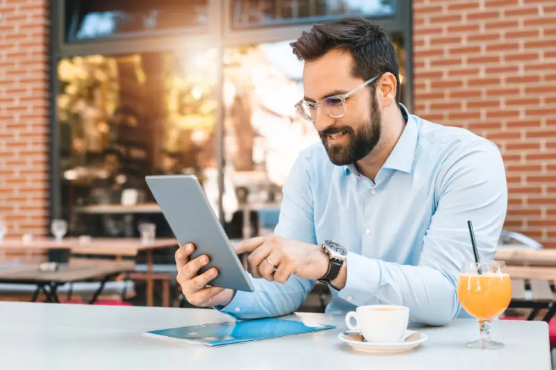 Businessman sitting outdoors and having a coffee while reading a blog on his silver tablet.
