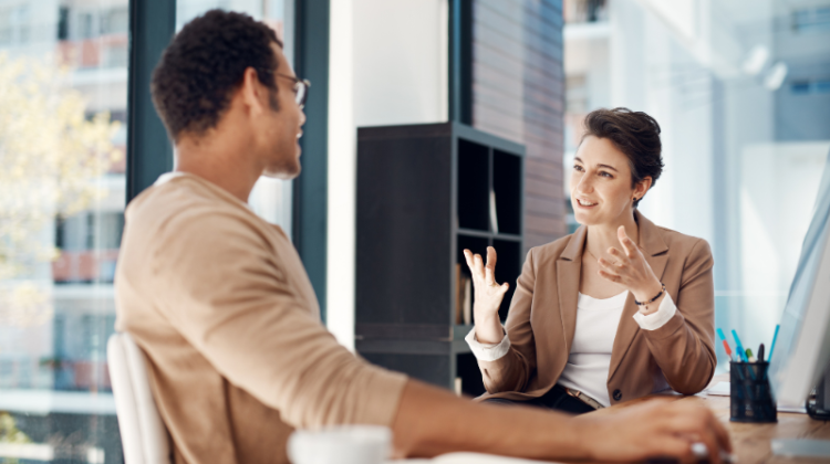 A woman gesturing during a one-to-one conversation with a man at an office desk.