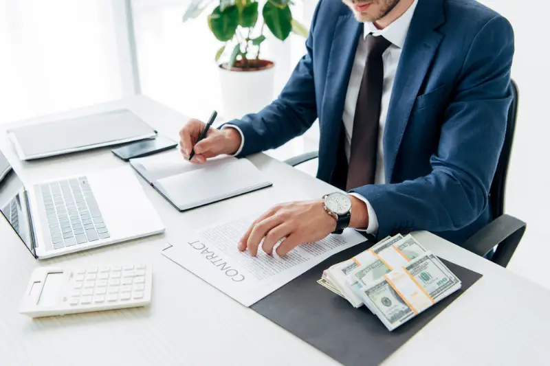 Cropped view of man in suit writing in notebook near money and laptop