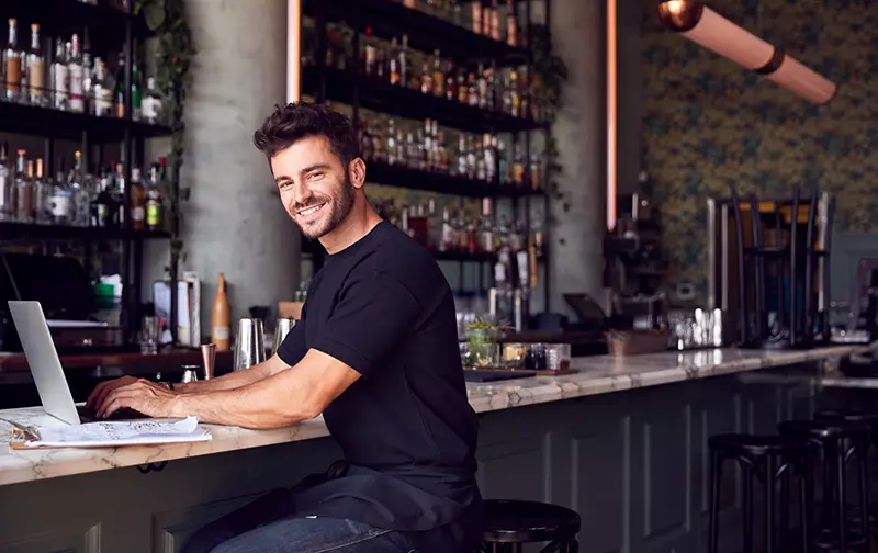 Male Owner Of Restaurant Bar Sitting At Counter Working On Laptop