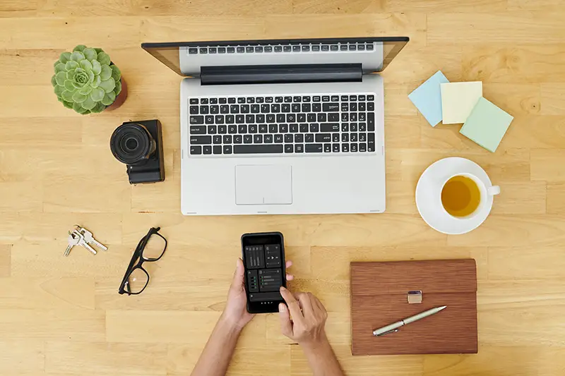 Image of woman sitting at wooden table in front of laptop computer and making a setting on her mobile phone