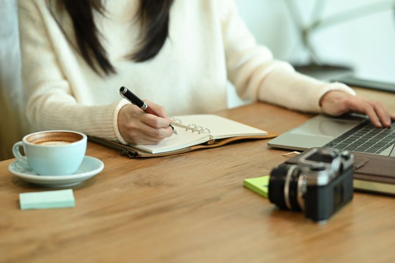 Young woman in sweater writing her diary on wooden table.