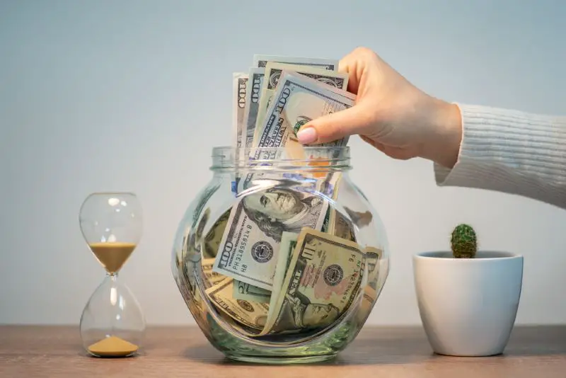 Female hand drops a bunch of dollar banknotes into a glass jar for savings