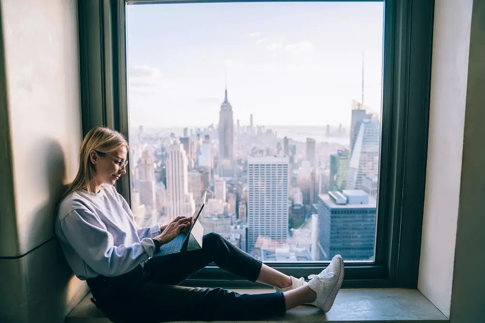 Remote worker using digital tablet while sitting on hotel window sill with view of New York cityscape