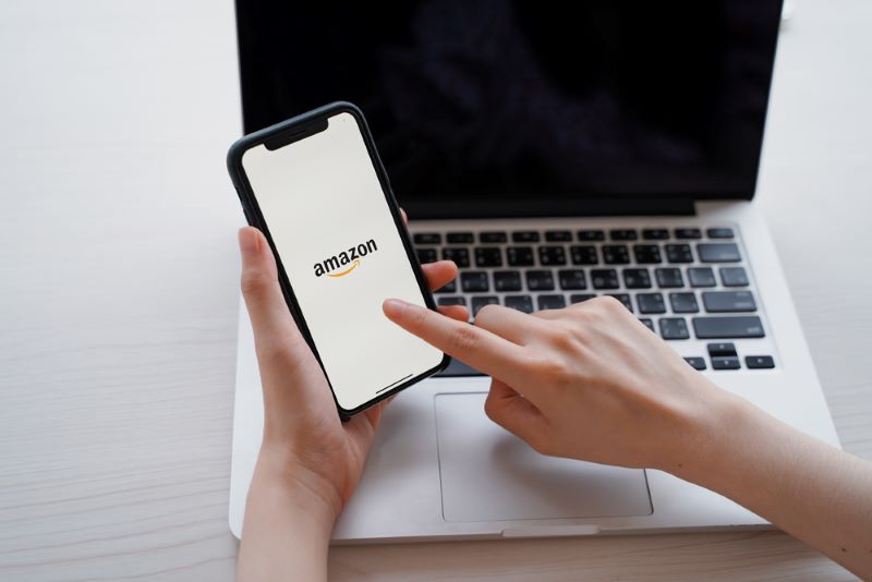woman using iPhone XS showing Amazon application on screen and laptop, Amazon is popular shopping online retailer