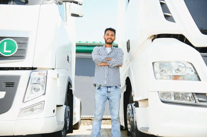 A confident Indian truck driver stood between two trucks parked in a car park.