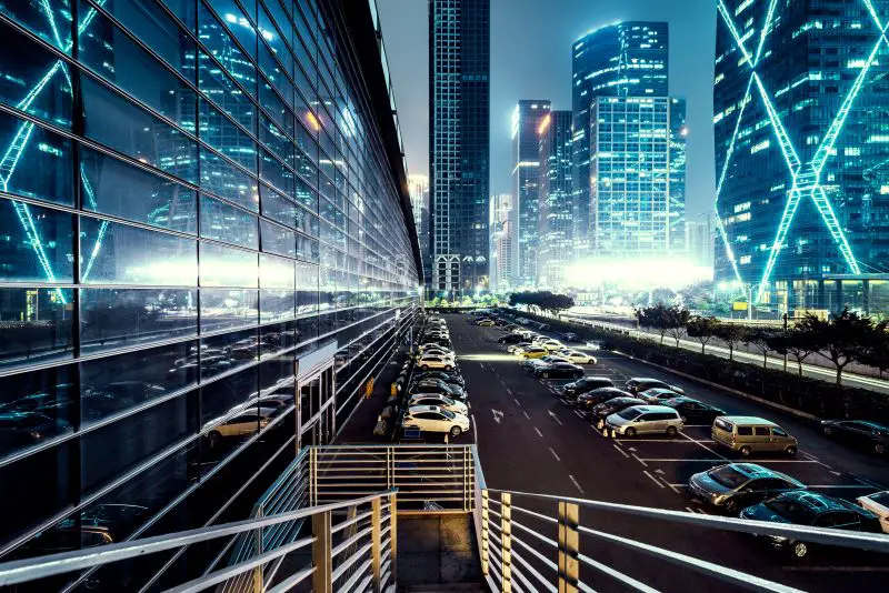 Modern architecture lit up at night next to a car park full of vehicles.