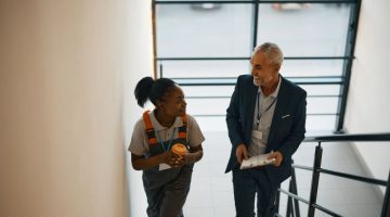 Happy mature manager with clipboard and black female worker talking while walking up the stairs