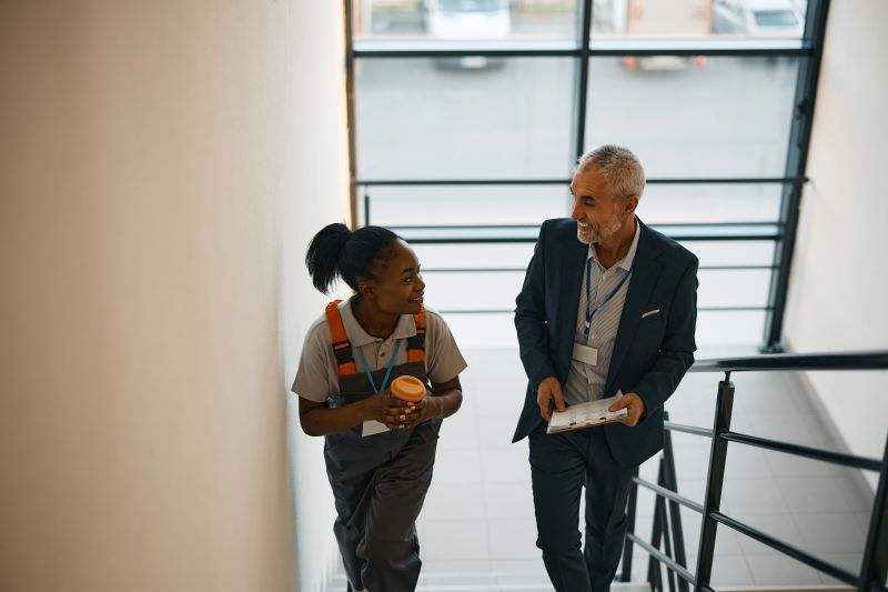 Happy mature manager with clipboard and black female worker talking while walking up the stairs