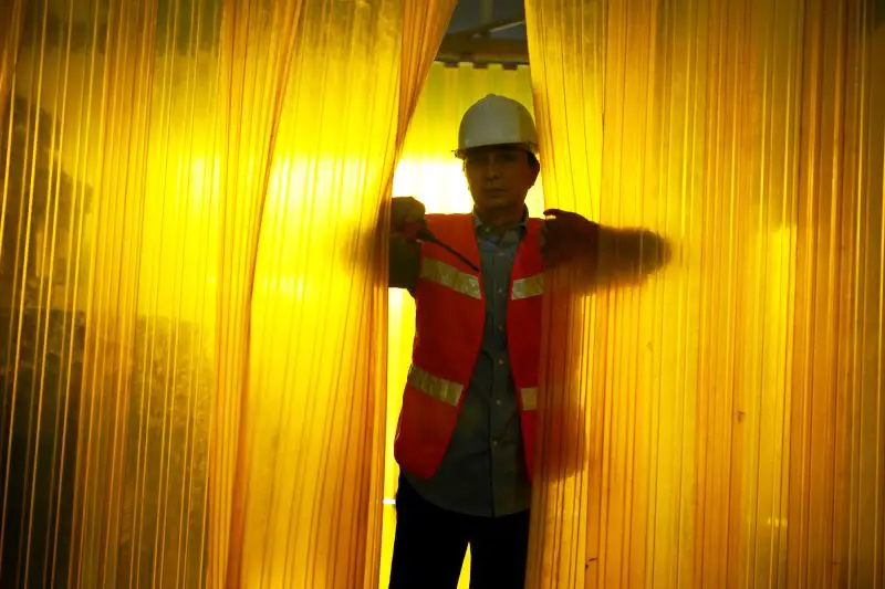 A worker wearing a hard hat is peering through a PVC curtain in a factory.